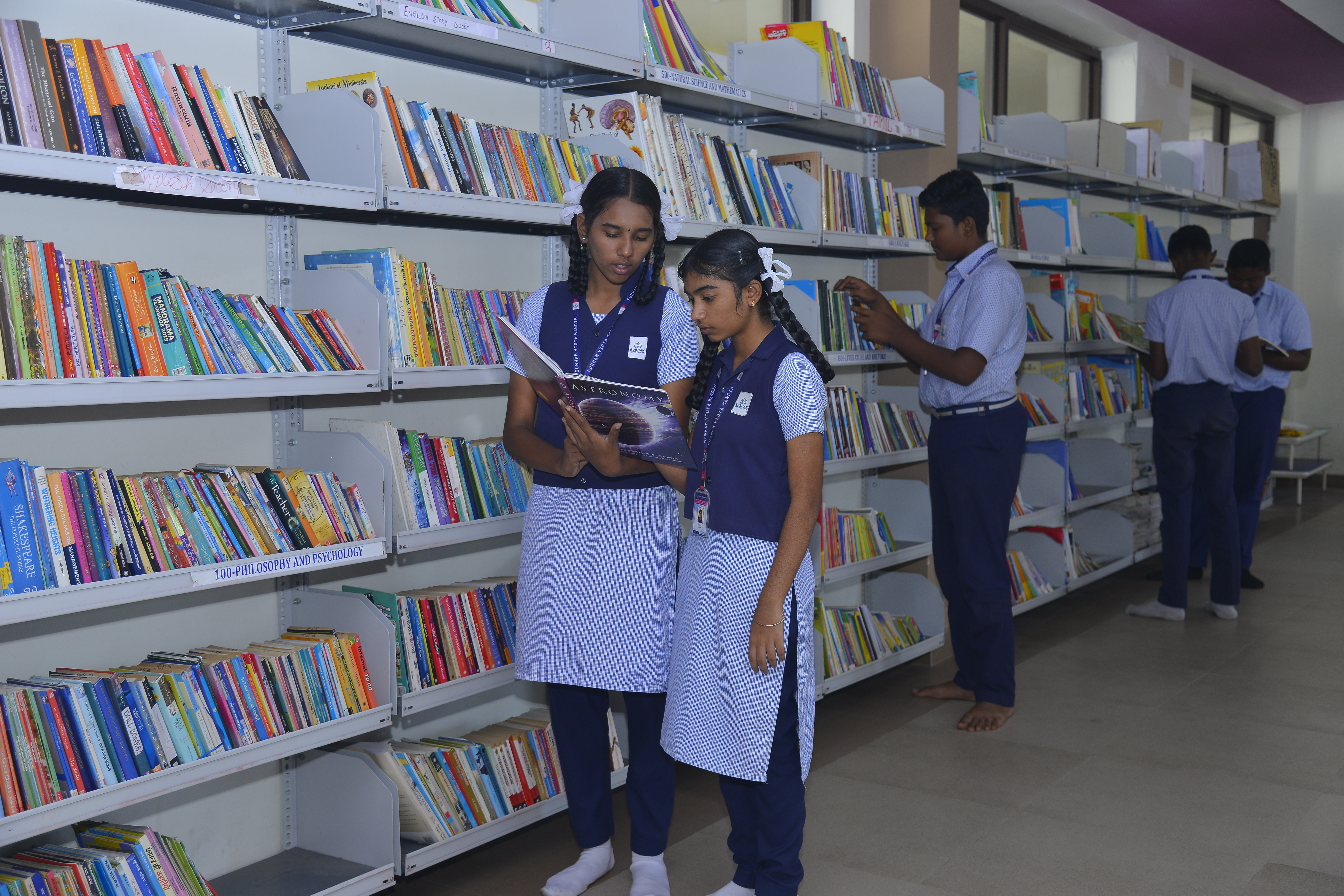 Inside view of School library with  books arranged on the shelves and three staff sitting on the table and three more sitting down and reading